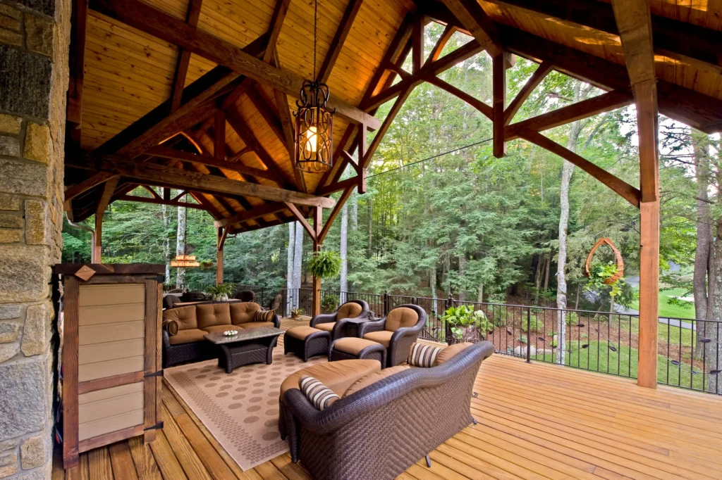 Rustic covered timber frame deck featuring high vaulted wood ceiling beams, cozy lounge seating, and forest views.